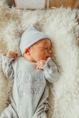 Sleeping newborn in grey 'Heaven Grace' onesie and hat on fluffy white blanket.