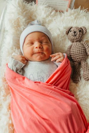 Peaceful newborn in grey outfit and hat wrapped in coral blanket with teddy bear.