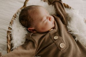 Sleeping newborn wearing brown knit outfit in cozy basket with white fluff.