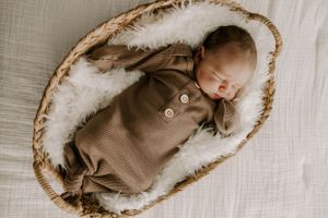 Newborn in brown buttoned outfit sleeps peacefully in rustic basket.