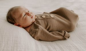 Sleeping newborn wearing cozy brown knit sweater on white bedding.