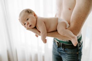 Parent holding naked newborn baby sideways against window light.