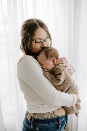 A tender moment as someone in glasses and white sweater cradles a baby wearing a brown knit outfit by white curtains.