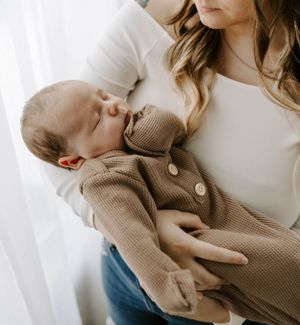 Someone in white holds a sleeping baby dressed in a cozy brown cardigan against a bright window.