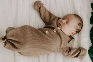 Sleeping baby wearing a tan knitted romper with buttons lies on white bedding.
