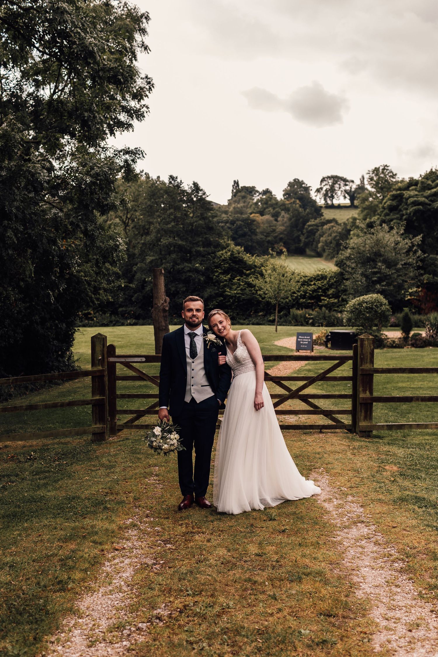 A newlywed pair poses in front of a rustic wooden gate on their wedding day in the countryside.