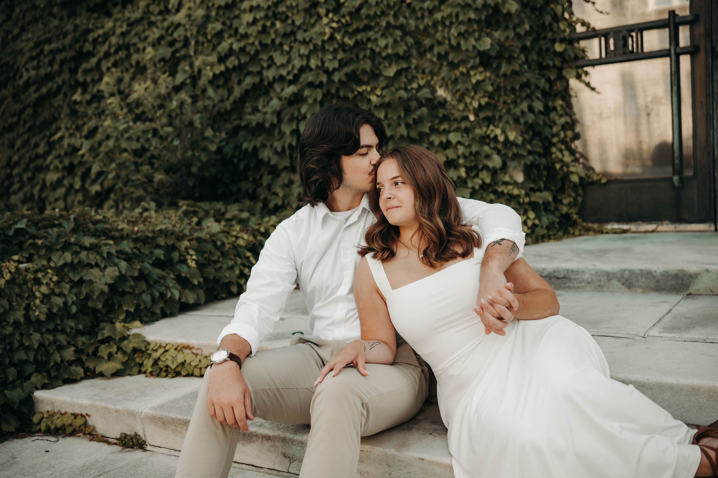 Couple sitting on stone steps embracing before ivy-covered wall in wedding attire.