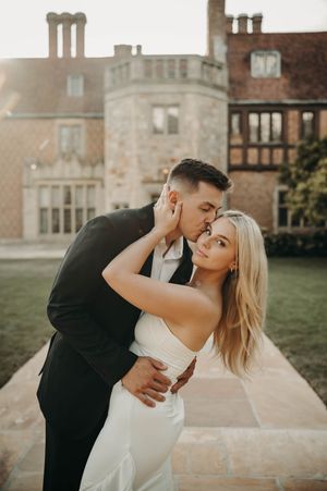 Romantic couple in wedding attire share a tender kiss outside a grand historic stone manor at sunset.