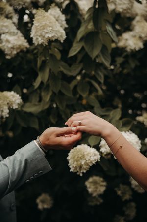 Close-up of couple holding hands with an engagement ring near white hydrangea flowers in a garden.