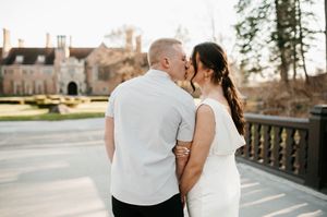Couple sharing romantic kiss on outdoor bridge near historic brick building at sunset.