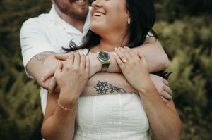 Couple embracing outdoors, woman in white dress with floral tattoo, man hugging her from behind, both smiling.