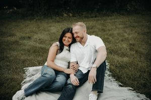A smiling couple sits together on a blanket in a grassy park, dressed casually in jeans and white tops.
