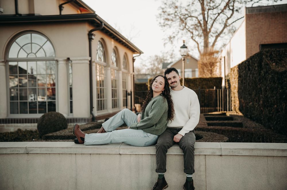 Casual couple sitting on a stone ledge outside a charming brick building at golden hour.