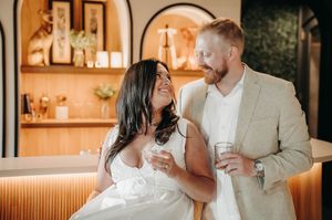 Smiling couple holding drinks at a modern bar, woman in white dress and man in tan blazer gazing at each other.