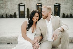 A smiling couple in elegant attire sitting together on stone steps outside a rustic building.