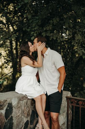 Couple shares a romantic kiss on a stone wall surrounded by lush green trees during an outdoor session.