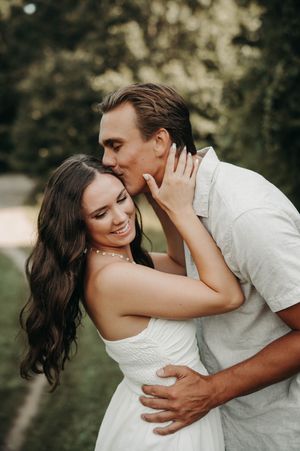 Man kisses woman on the cheek while embracing her from behind in a sunny outdoor engagement photo.