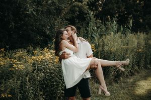 Man lifts and kisses woman in a white dress amid vibrant green foliage in a romantic outdoor portrait.