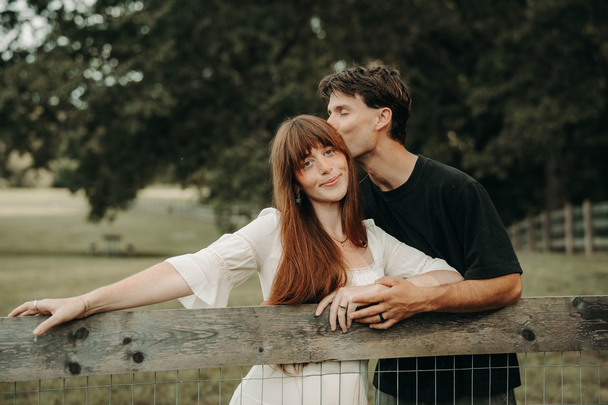 Romantic couple posing by a rustic wooden fence in a lush green park, man kissing woman's cheek tenderly.