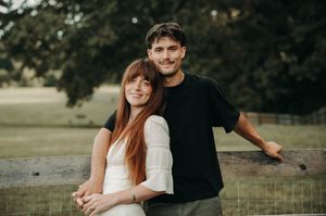 A smiling couple poses outdoors by a wooden fence with lush green trees and fields in the background.
