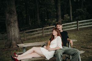 Couple relaxing on a wooden park bench surrounded by lush green trees and a rustic fence.