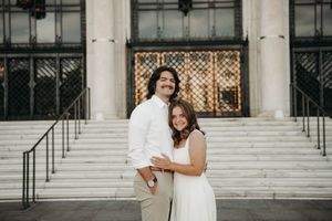 Couple embracing and smiling in front of a grand building with steps, wearing white outfits for an engagement photo.