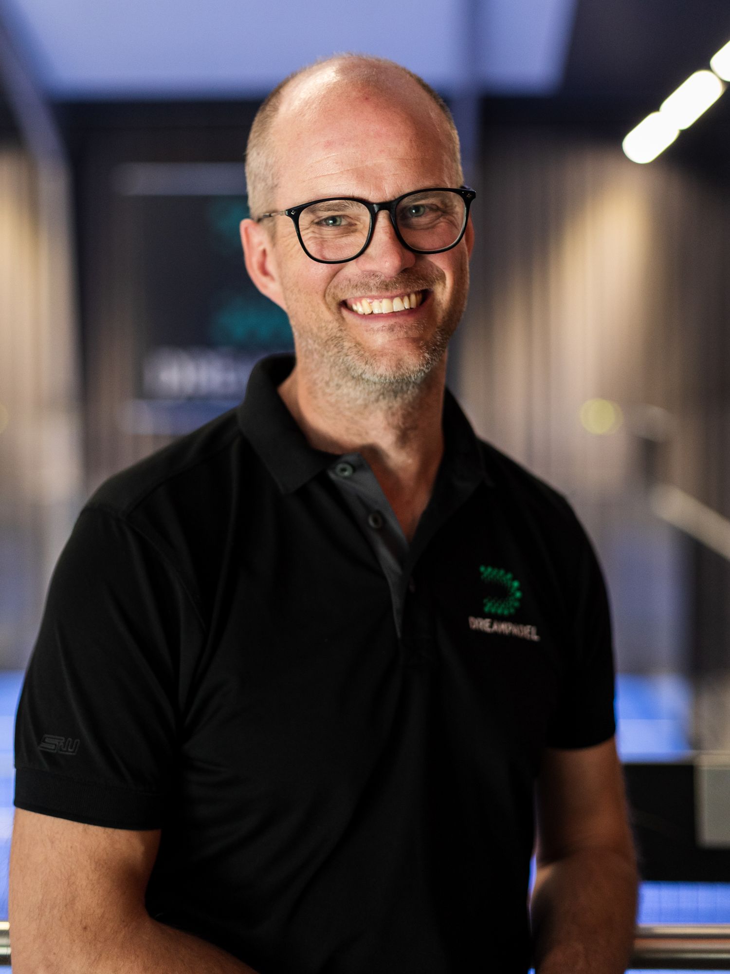 Professional in black polo shirt and glasses smiling in modern office setting.