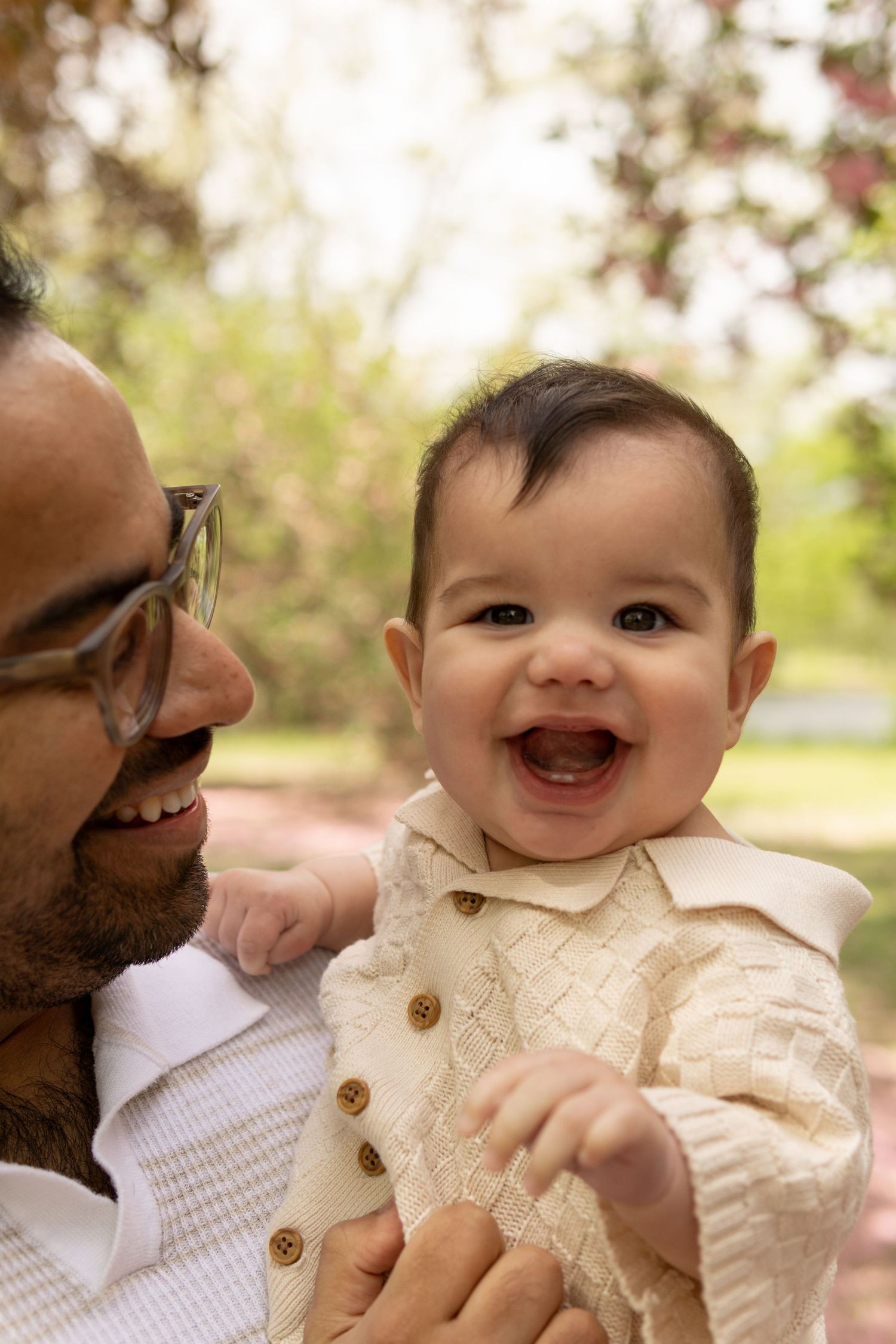 A baby wearing a cream-colored outfit smiles brightly during a springtime outdoor portrait session.