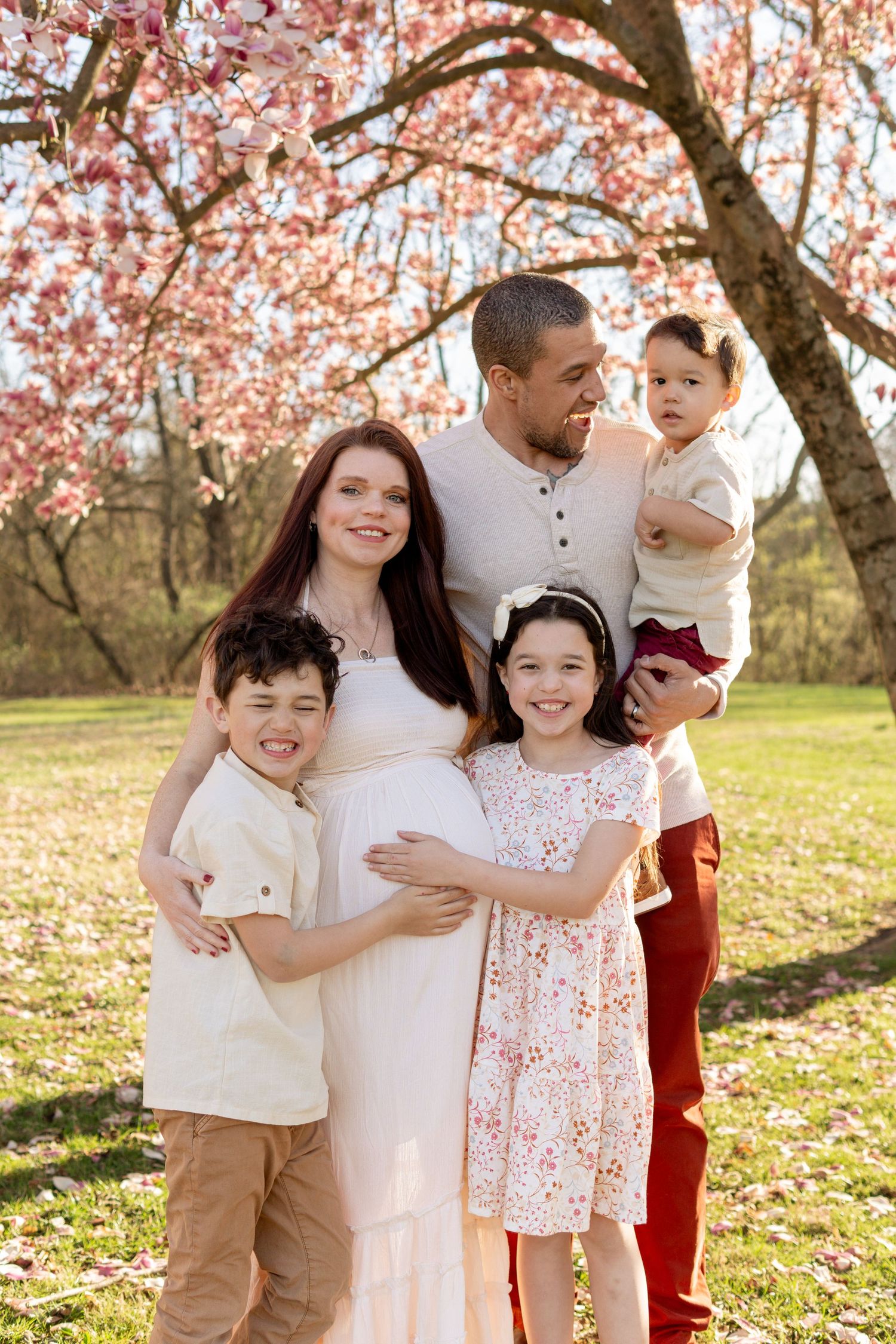 A family stands together under blooming cherry blossom trees in spring, with the mother wearing a white maternity dress.