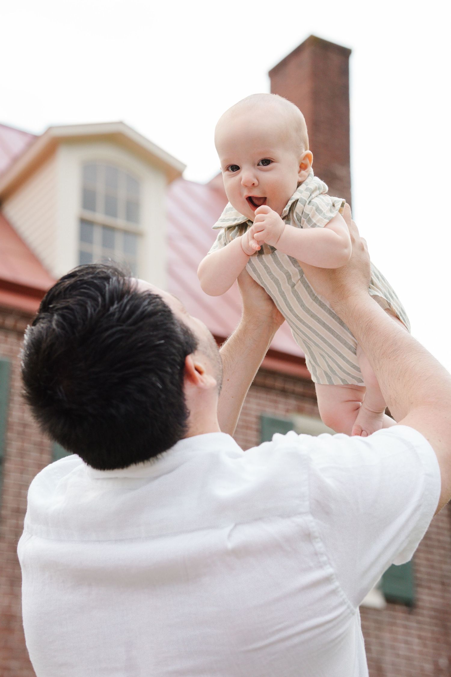 A baby in a striped romper being lifted up against a brick house exterior.