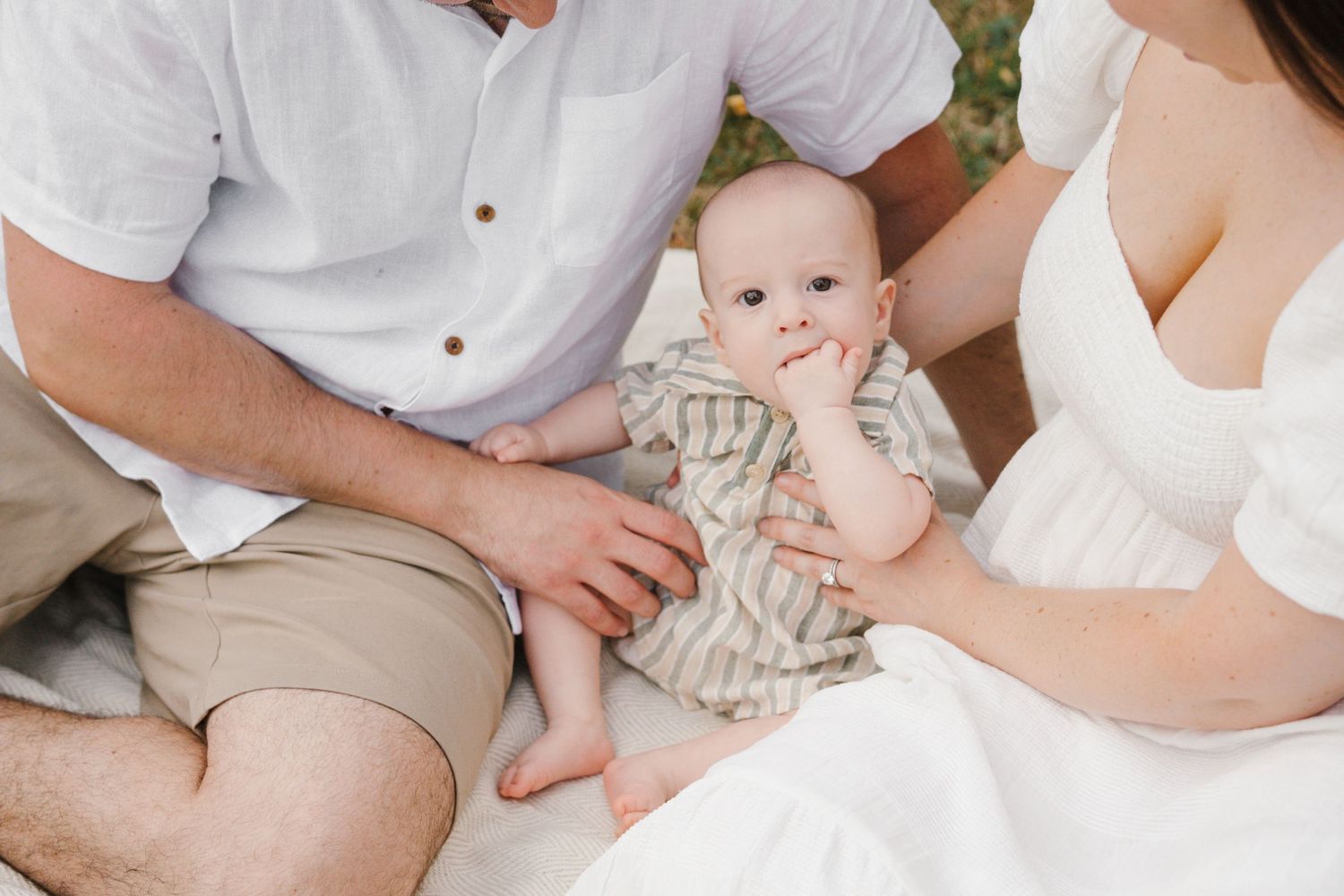 A baby in striped clothing sitting with adults on a white blanket outdoors.