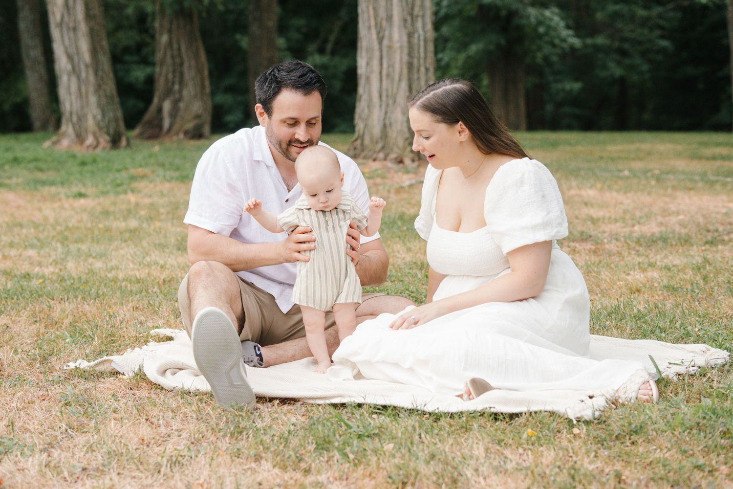 A couple sits on a white blanket having a picnic with their baby in a wooded setting.