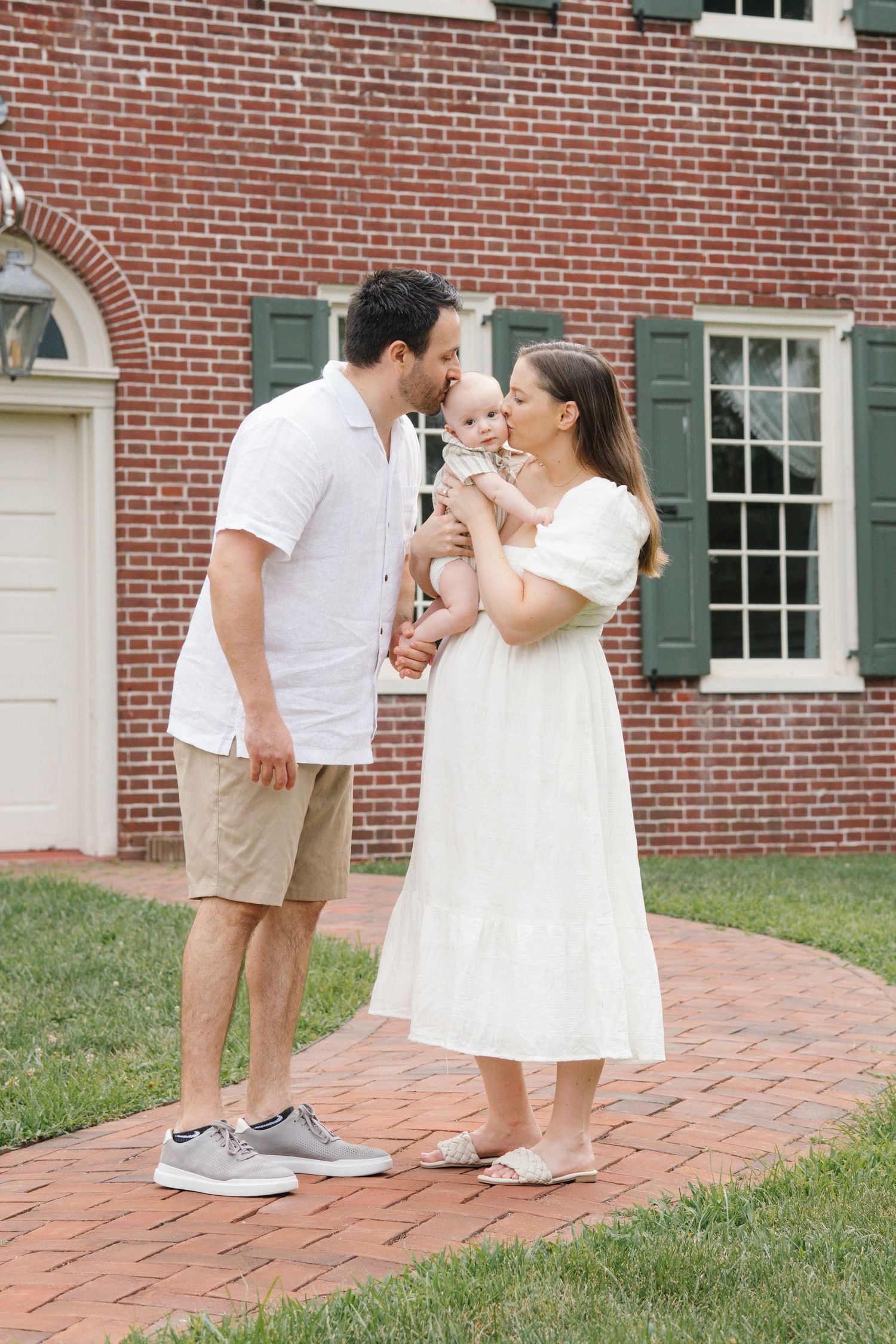 A couple in white summer clothing shares an intimate moment with their baby on a brick patio.