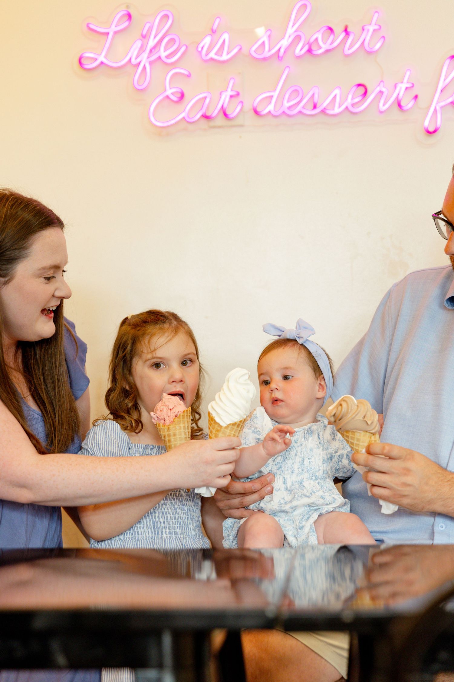 A family enjoying ice cream cones together under a pink neon sign that reads 'Life is short eat dessert'.