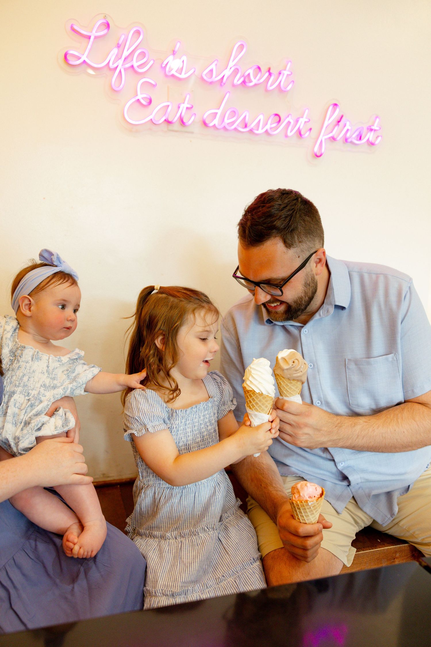 A family enjoying ice cream cones together under a neon sign that reads 'Life is short, Eat dessert first'.