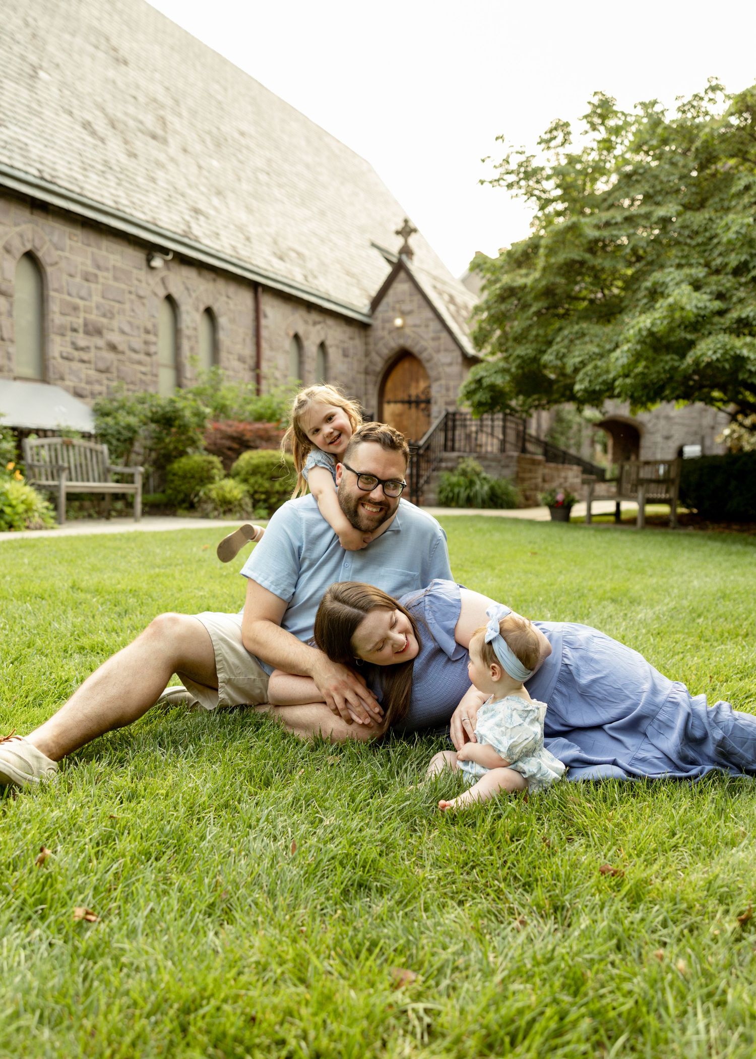 A couple lies together on lush green grass near a stone church on a sunny summer day.