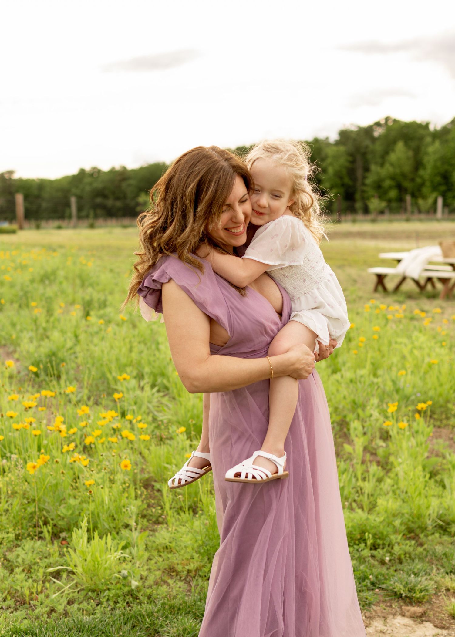 Two people hugging joyfully in a sunny meadow filled with vibrant wildflowers and picnic tables in the background.