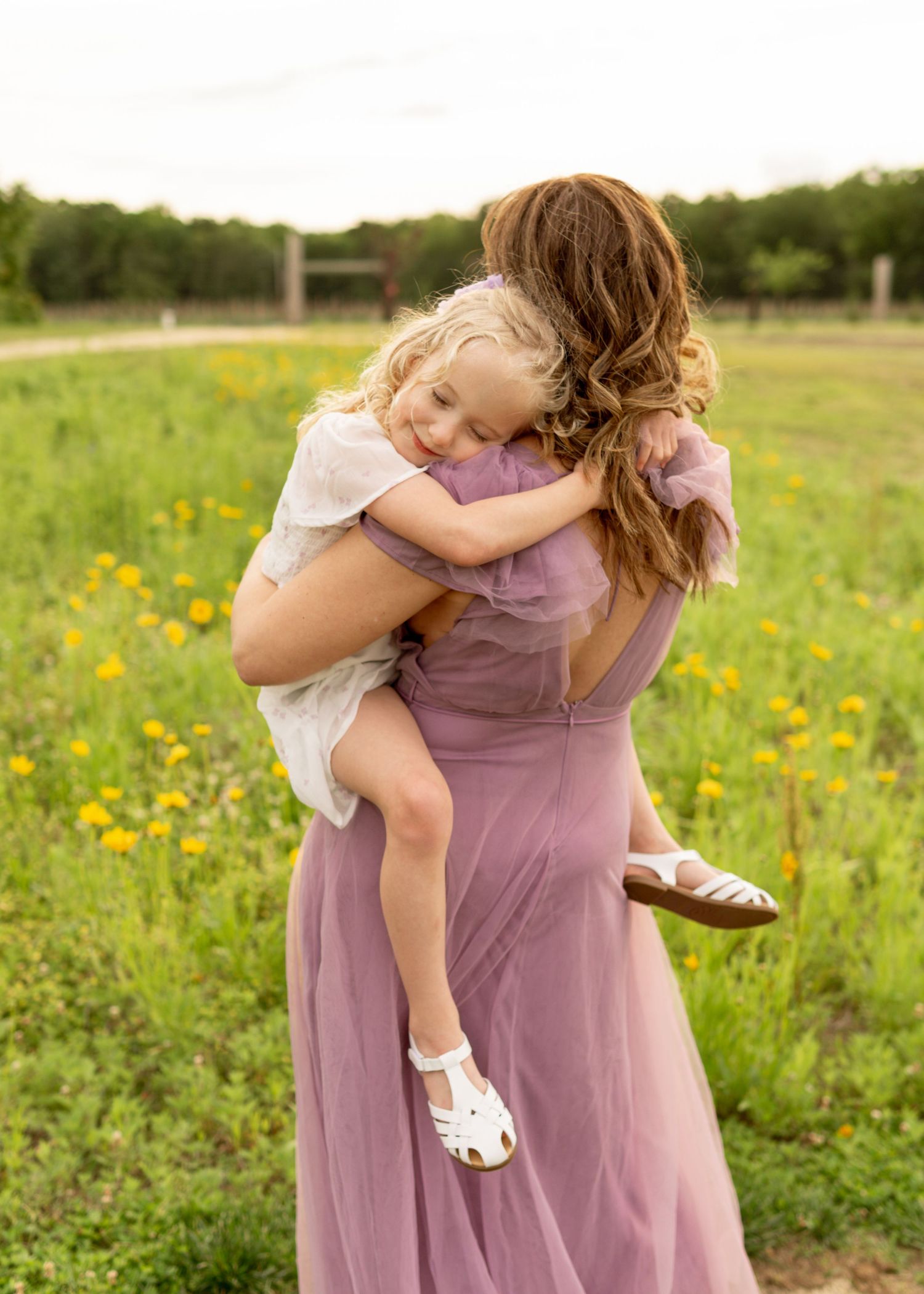 Mother and child embracing in a field of yellow wildflowers on a sunny afternoon wearing matching mauve outfits.
