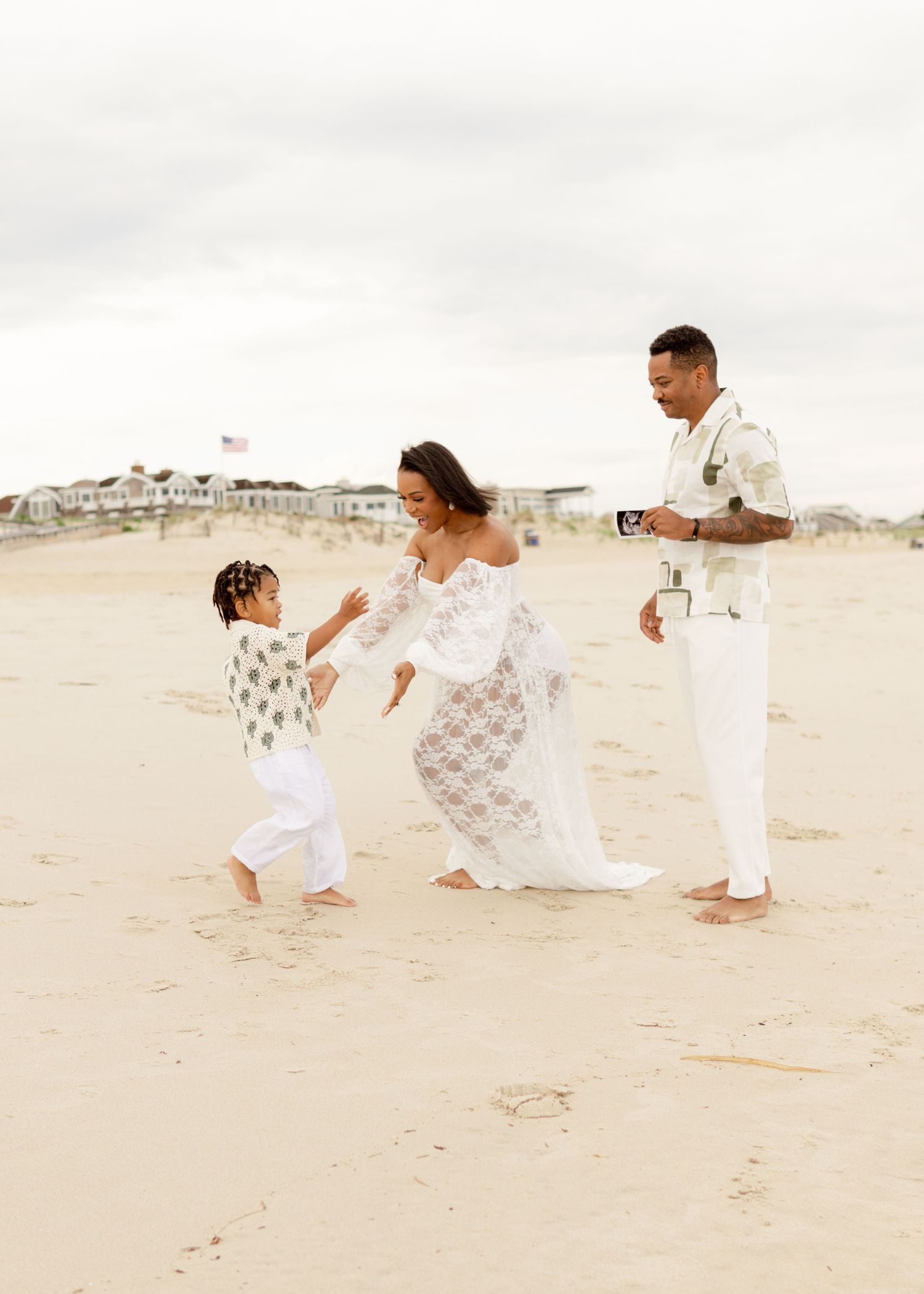 A happy family in coordinated white outfits dances together on a sandy beach with houses in the background.