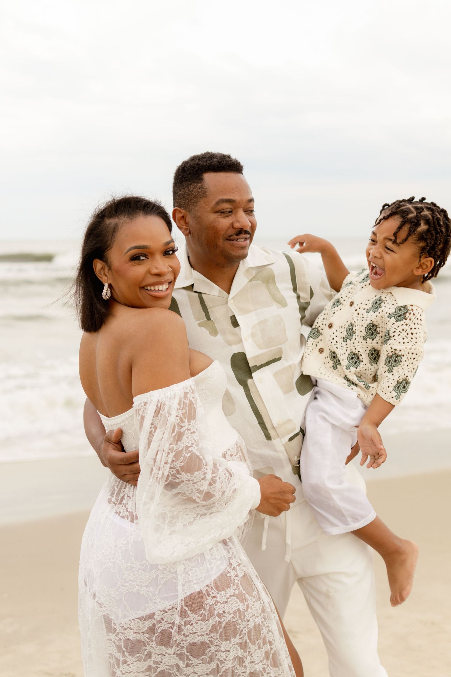 A family shares a joyful moment together on a sunny beach, wearing elegant white and cream colored outfits.