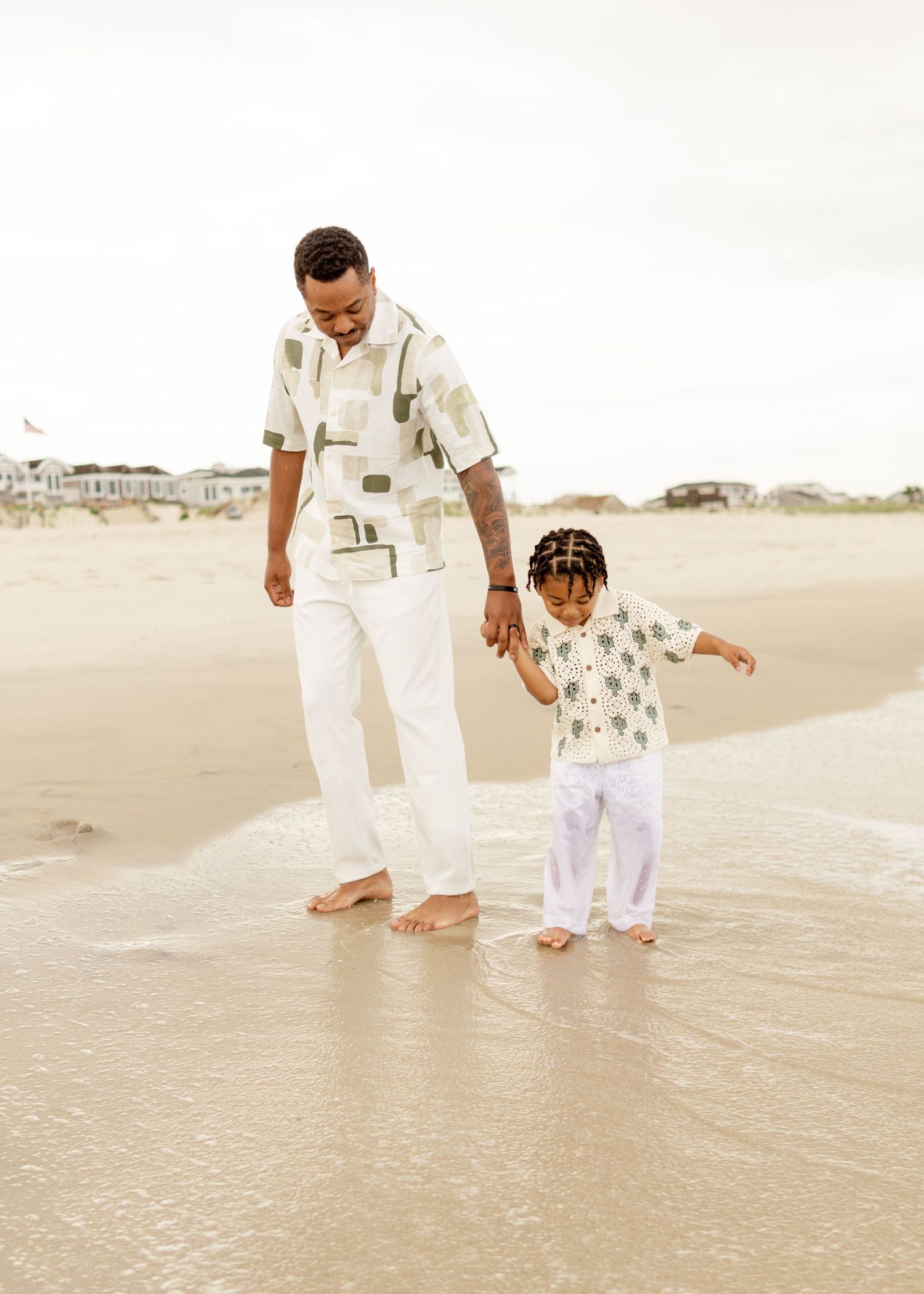 A parent and child walk barefoot on the beach wearing matching white outfits on a cloudy day.