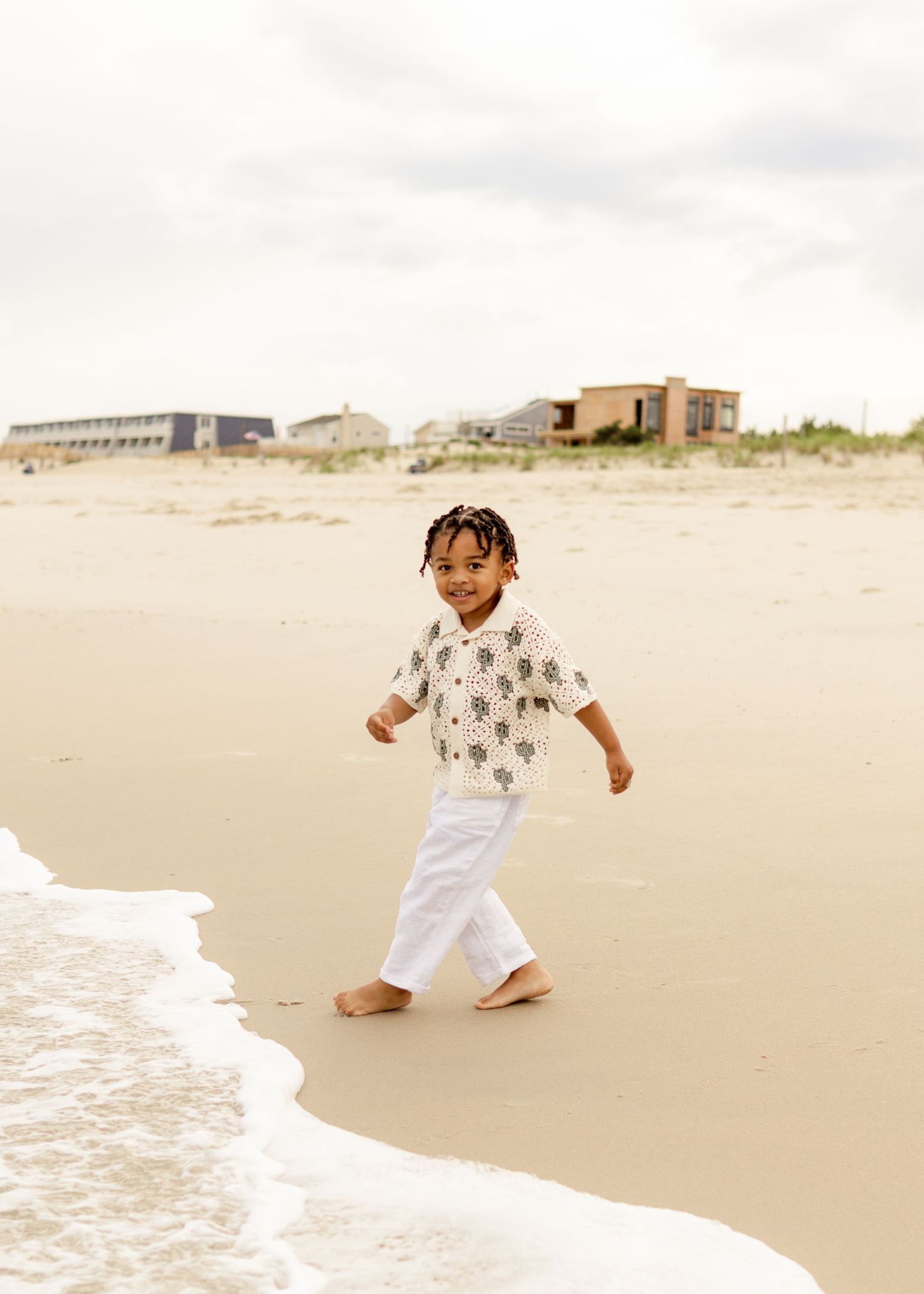 A child wearing a patterned shirt and white pants plays joyfully at the edge of ocean waves on a sandy beach.