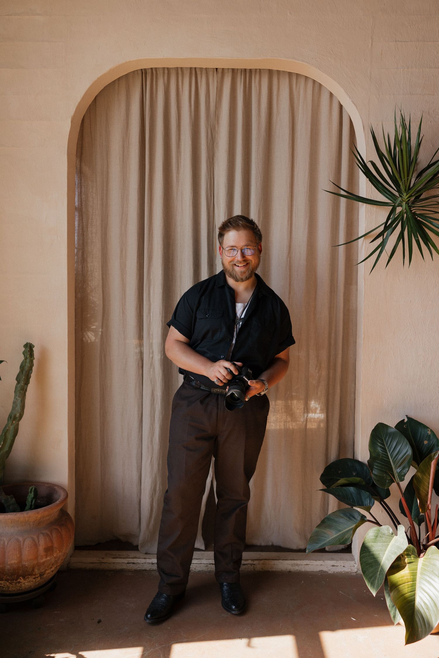 A photographer in casual attire poses with camera equipment against an arched doorway with indoor plants.