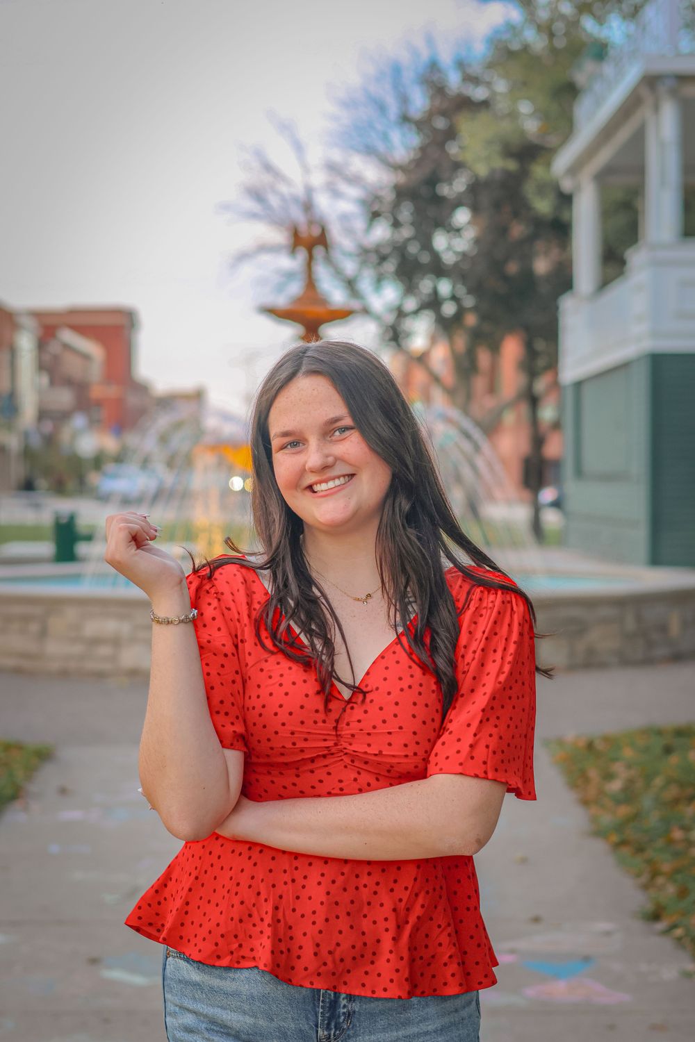 A person in a red patterned top poses playfully on a sidewalk with autumn trees and buildings in background.
