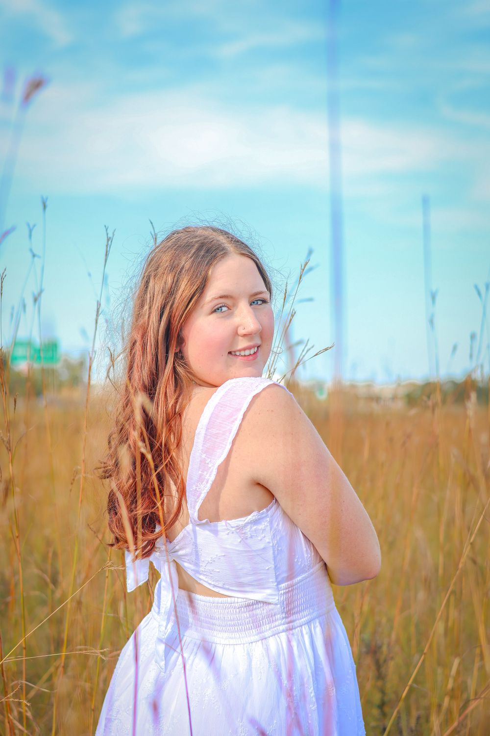 Woman in white dress poses gracefully in a field of tall grass against a blue sky.