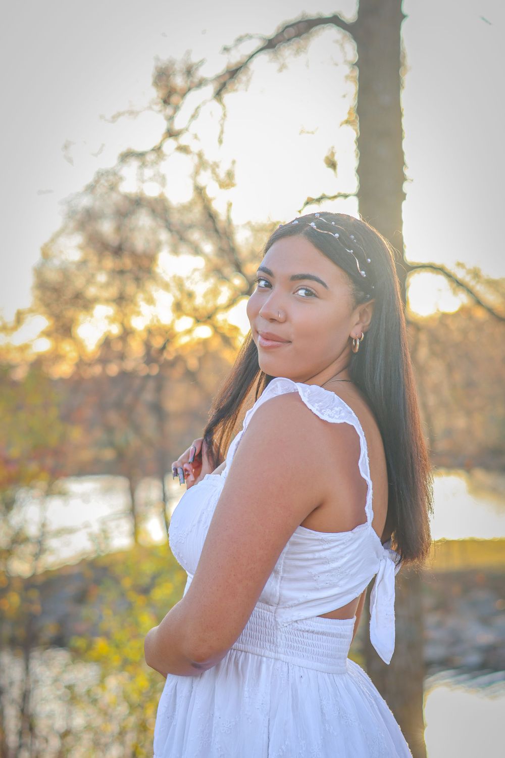 A woman in white dress poses against sunset with water and trees in background.