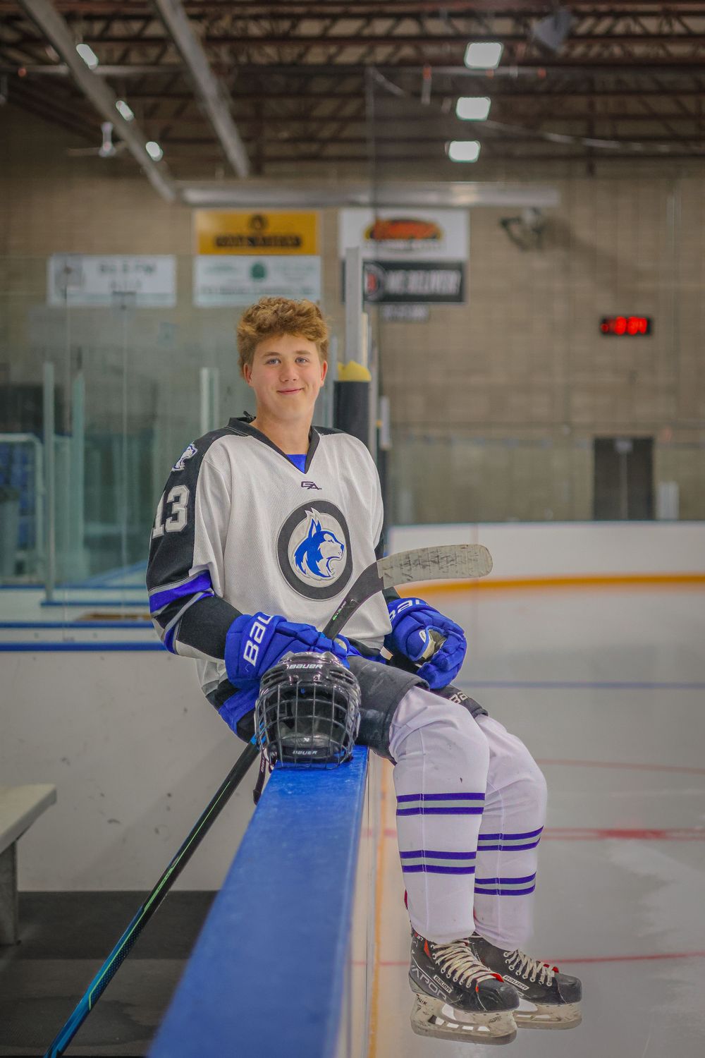 Hockey player rests on boards at ice rink wearing white and blue uniform.