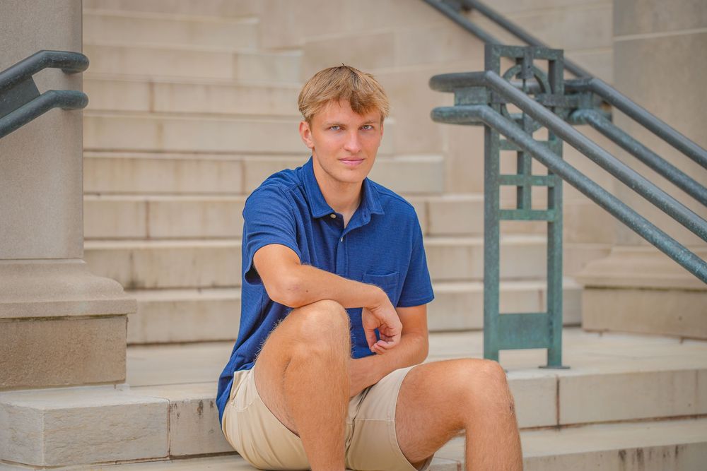 A person in a blue polo shirt and khaki shorts sits on concrete steps in front of a building.