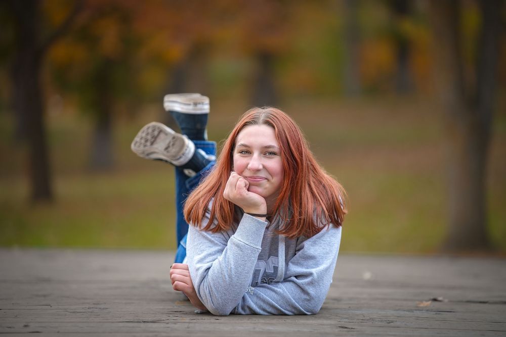 A person with red hair lies on wooden deck planks while holding a camera in an outdoor setting.