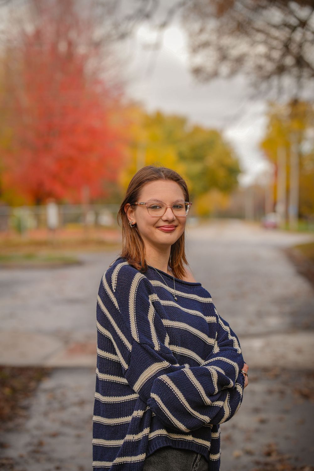 A person in a striped navy sweater poses outdoors on an autumn day with colorful fall foliage in the background.